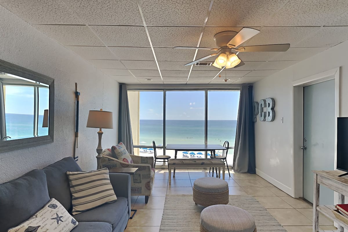 A spacious living room with a large window overlooking the beach, a ceiling fan, and a TV on a side table.