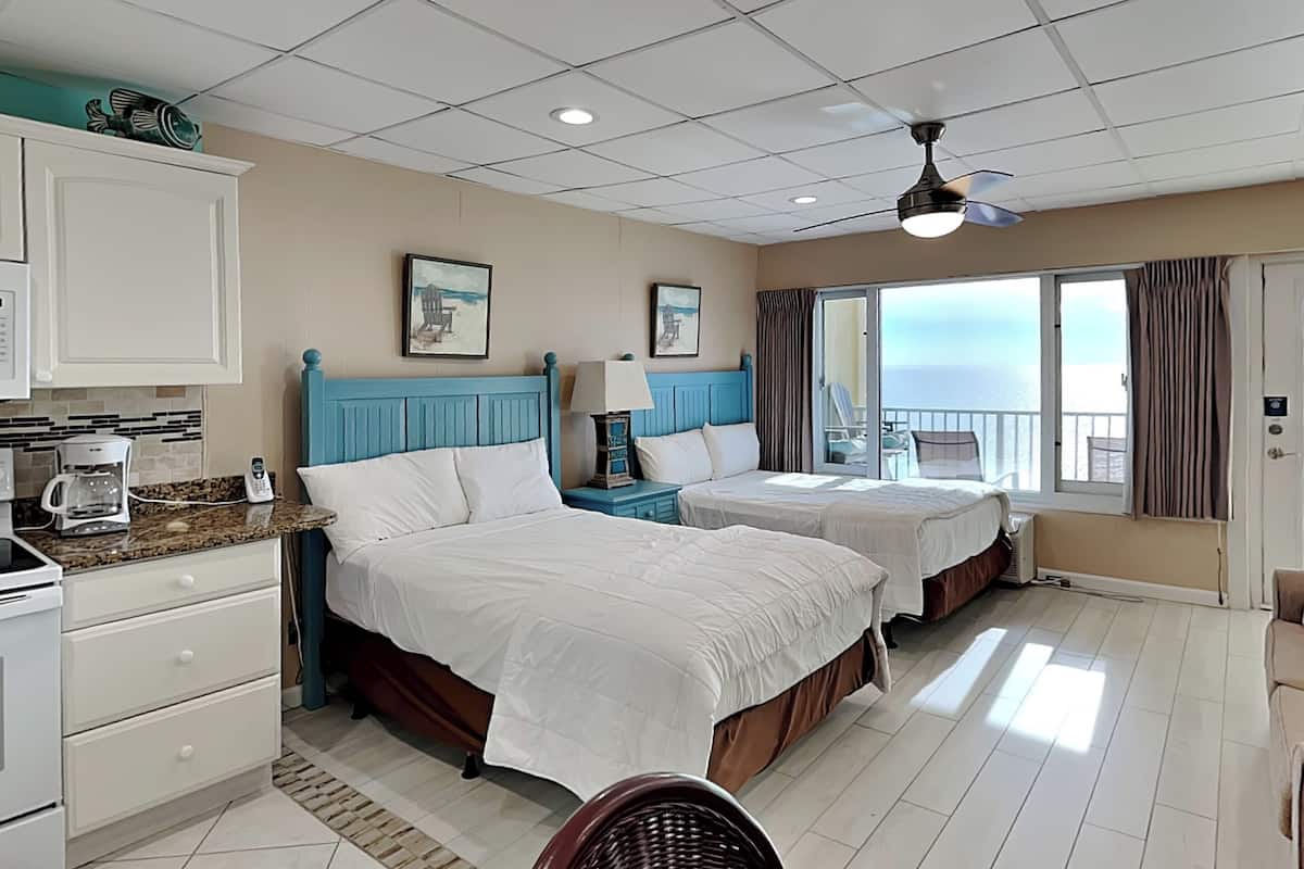 A hotel room with two beds, a kitchenette, a balcony with a view of the sea, and a ceiling fan.