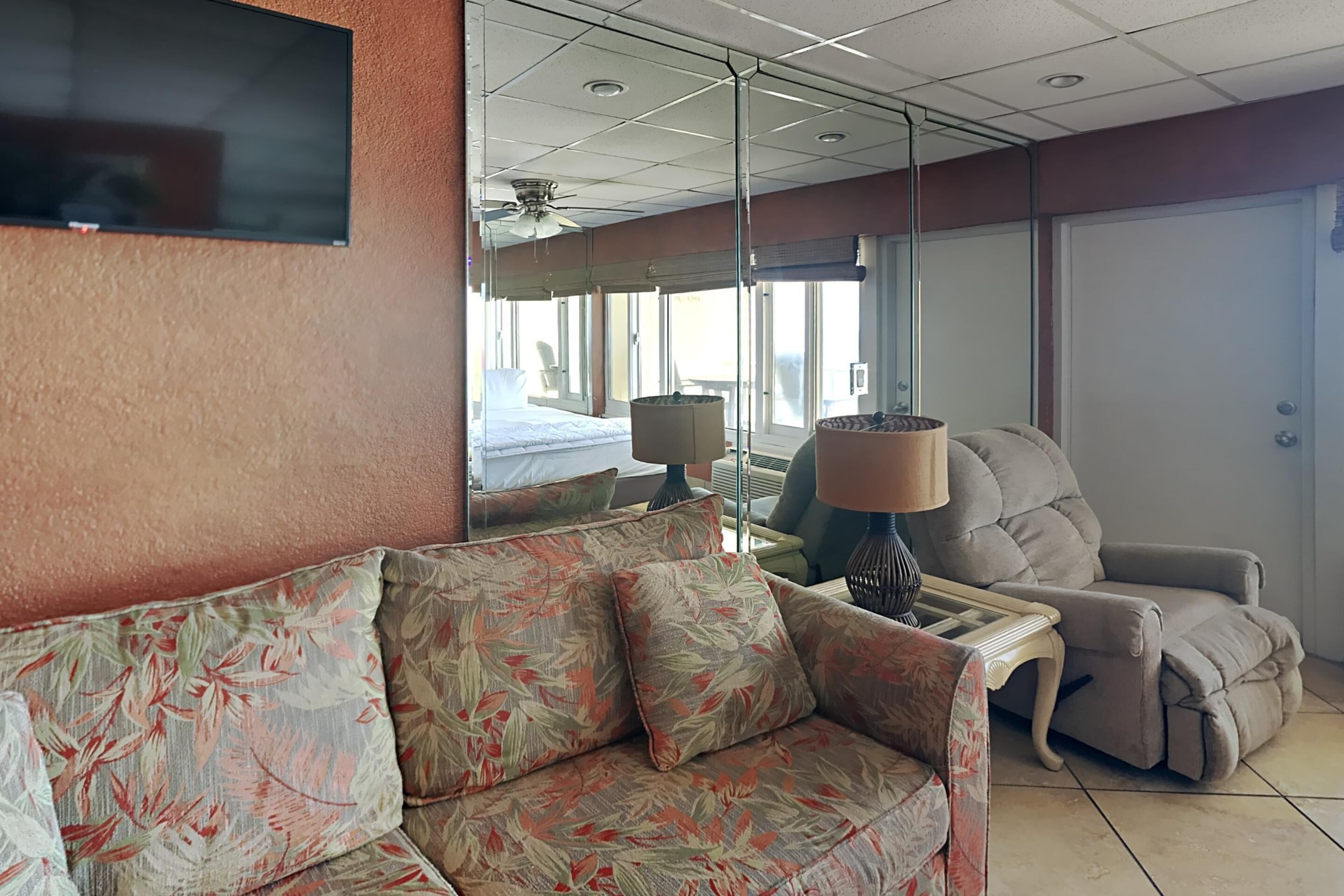 A hotel room with a sofa, a recliner, a bedside table with a lamp, a bed, and a large window.