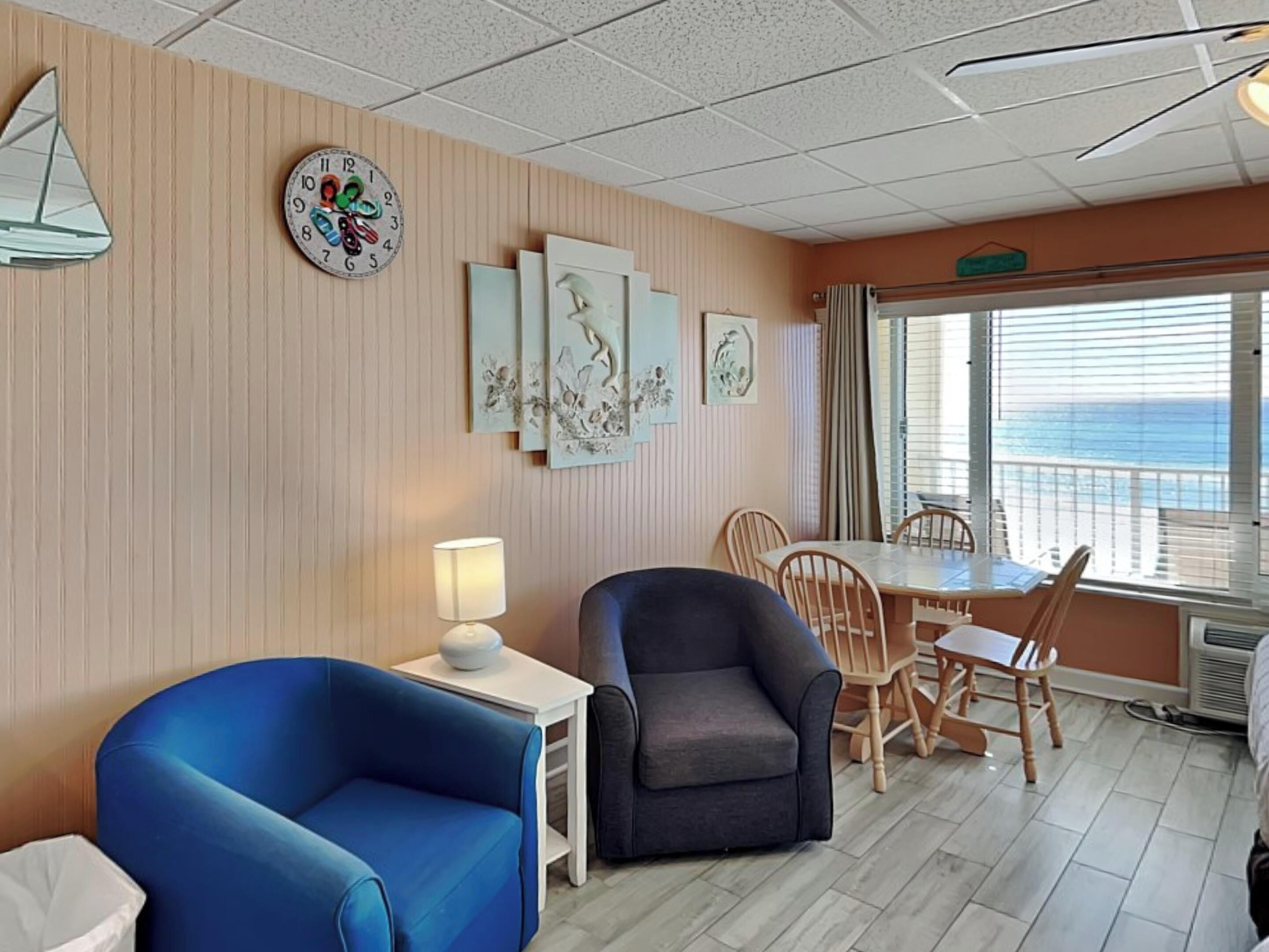 A room with a blue armchair, a grey armchair, a small table with a lamp, and a dining area with wooden chairs.