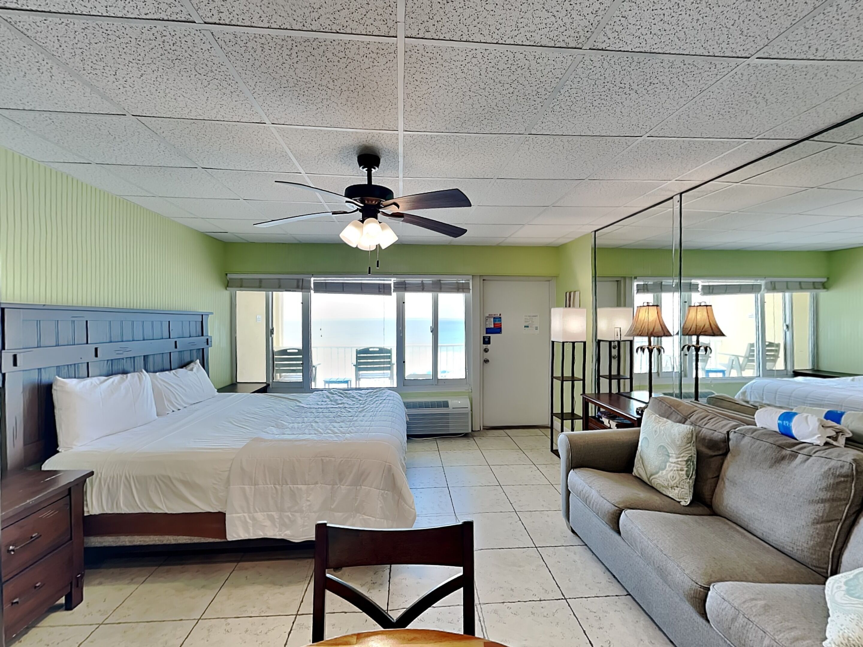 A hotel room with a bed, a sofa, a chair, a ceiling fan, and a view of the ocean through the balcony doors.
