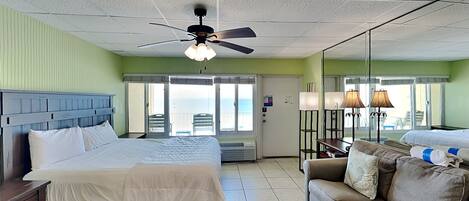 A hotel room with a bed, a sofa, a chair, a ceiling fan, and a view of the ocean through the balcony doors.
