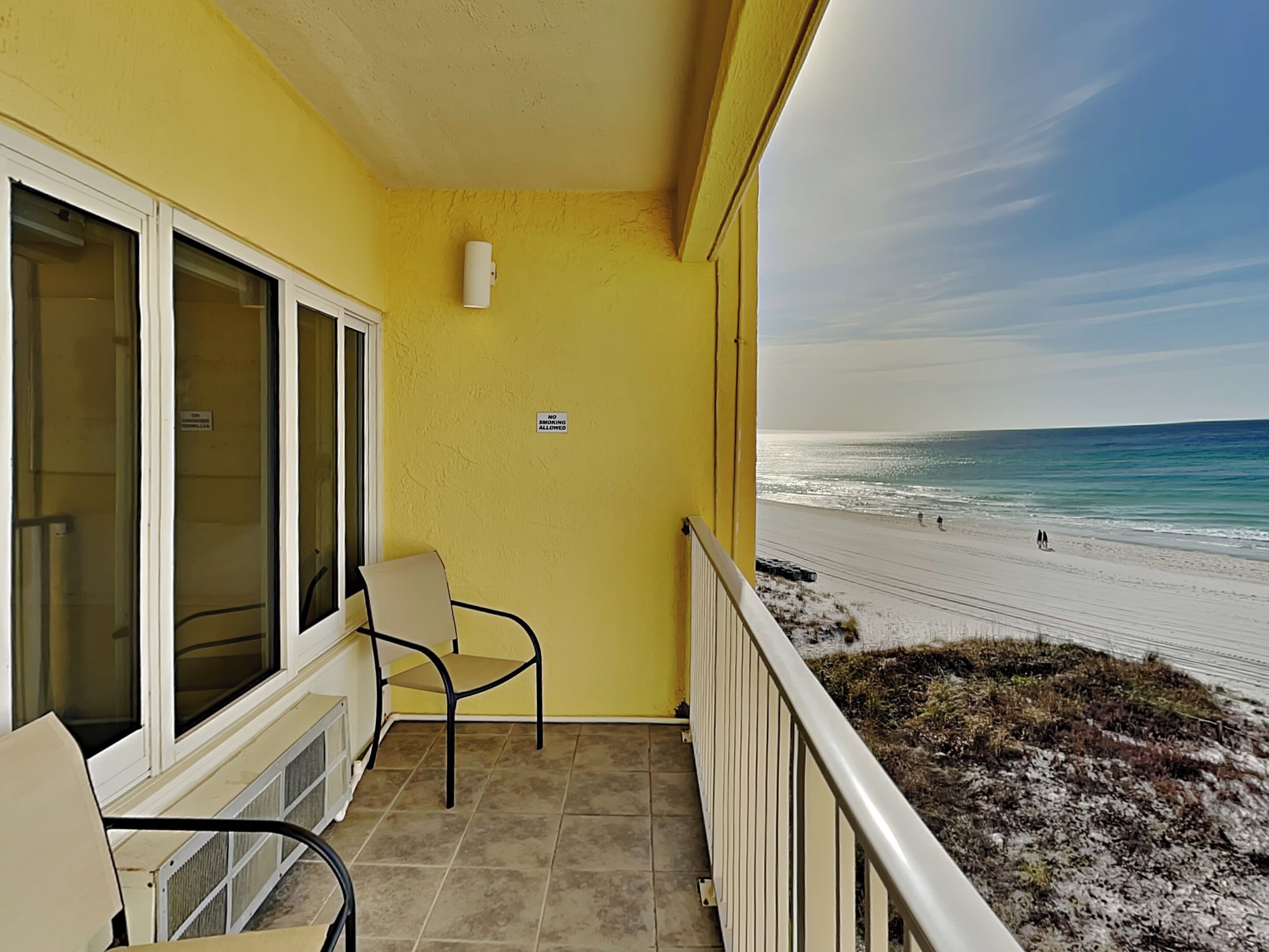 Un balcone con pareti gialle, una sedia e vista su una spiaggia con persone che passeggiano.