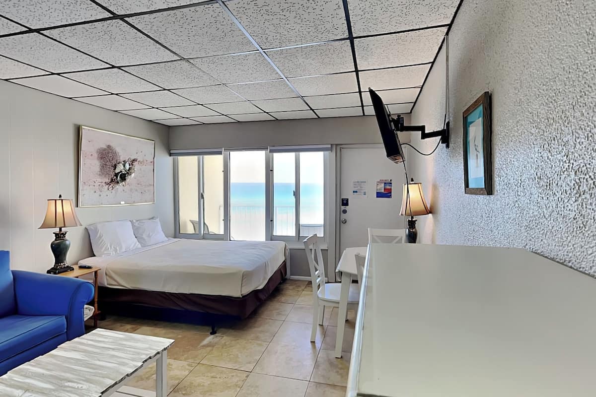 A hotel room with a bed, a blue sofa, a small table, a desk, and a view of the ocean through large windows.