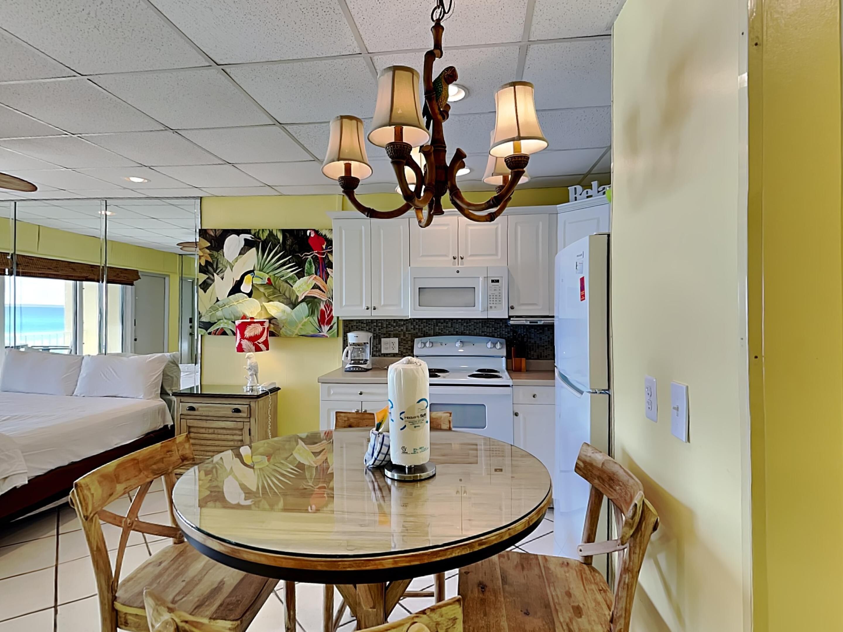 A hotel room with a bed, a round dining table with chairs, a kitchen area with a stove and microwave, and a ceiling with a chandelier.