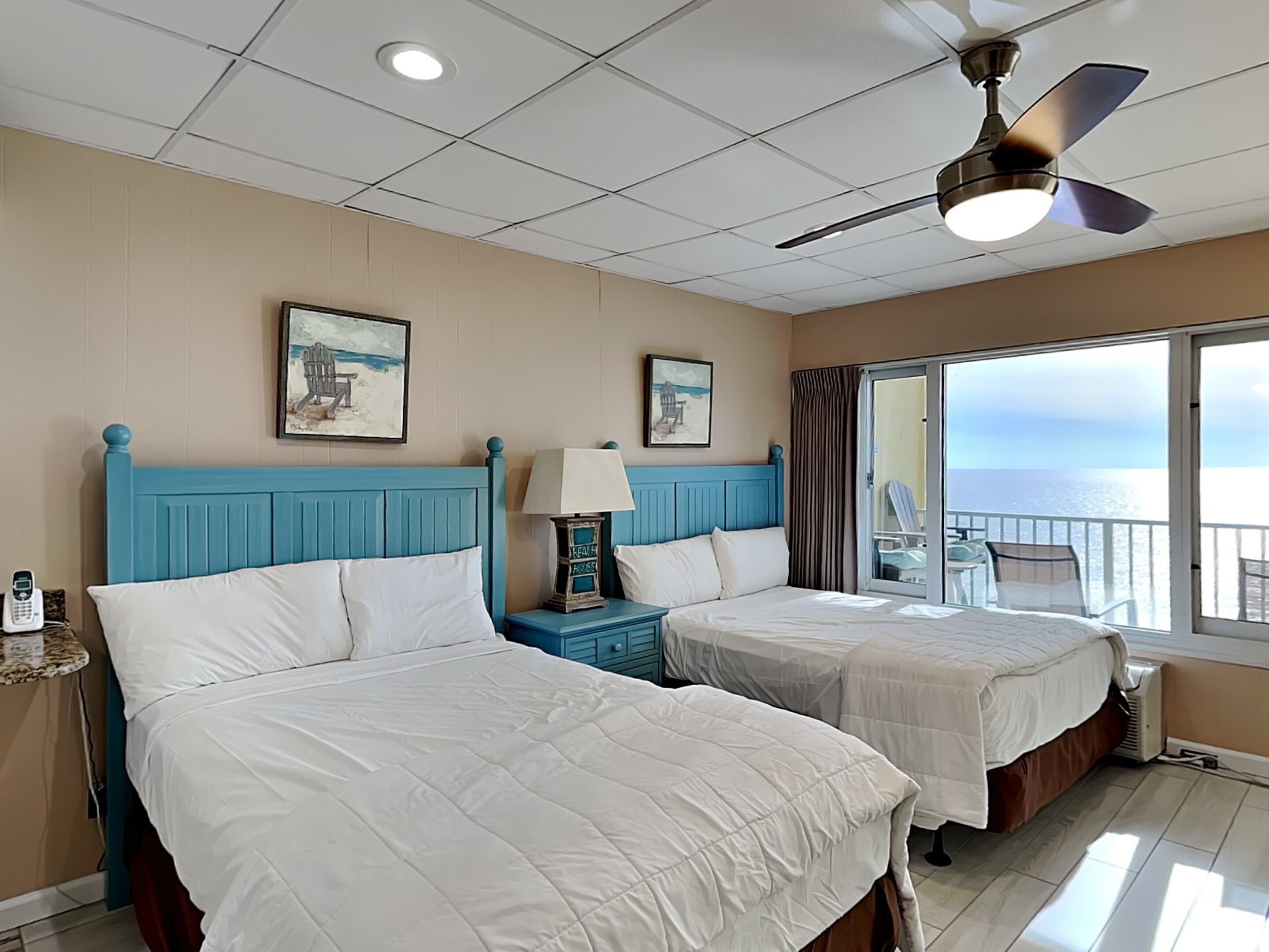 A hotel room with two beds, a ceiling fan, and a balcony with a view of the sea.