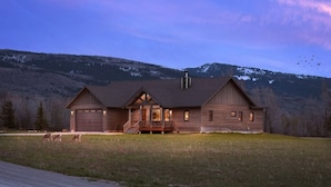 A two-story log cabin with a front porch and a garage, set against a mountainous backdrop. - Teton Time 4BR Home  - Hot Tub (Driggs)
