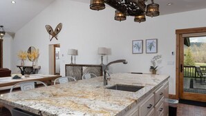A kitchen with a granite countertop, stainless steel sink, and white cabinetry. There is a dining area with a table and chairs, and a view of the outdoors through a glass door. - Teton Time 4BR Home  - Hot Tub (Driggs)