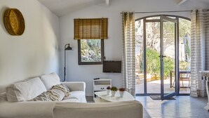 Un salon moderne avec un canapé blanc, une petite table blanche, un téléviseur et une porte-fenêtre coulissante donnant sur un espace extérieur.