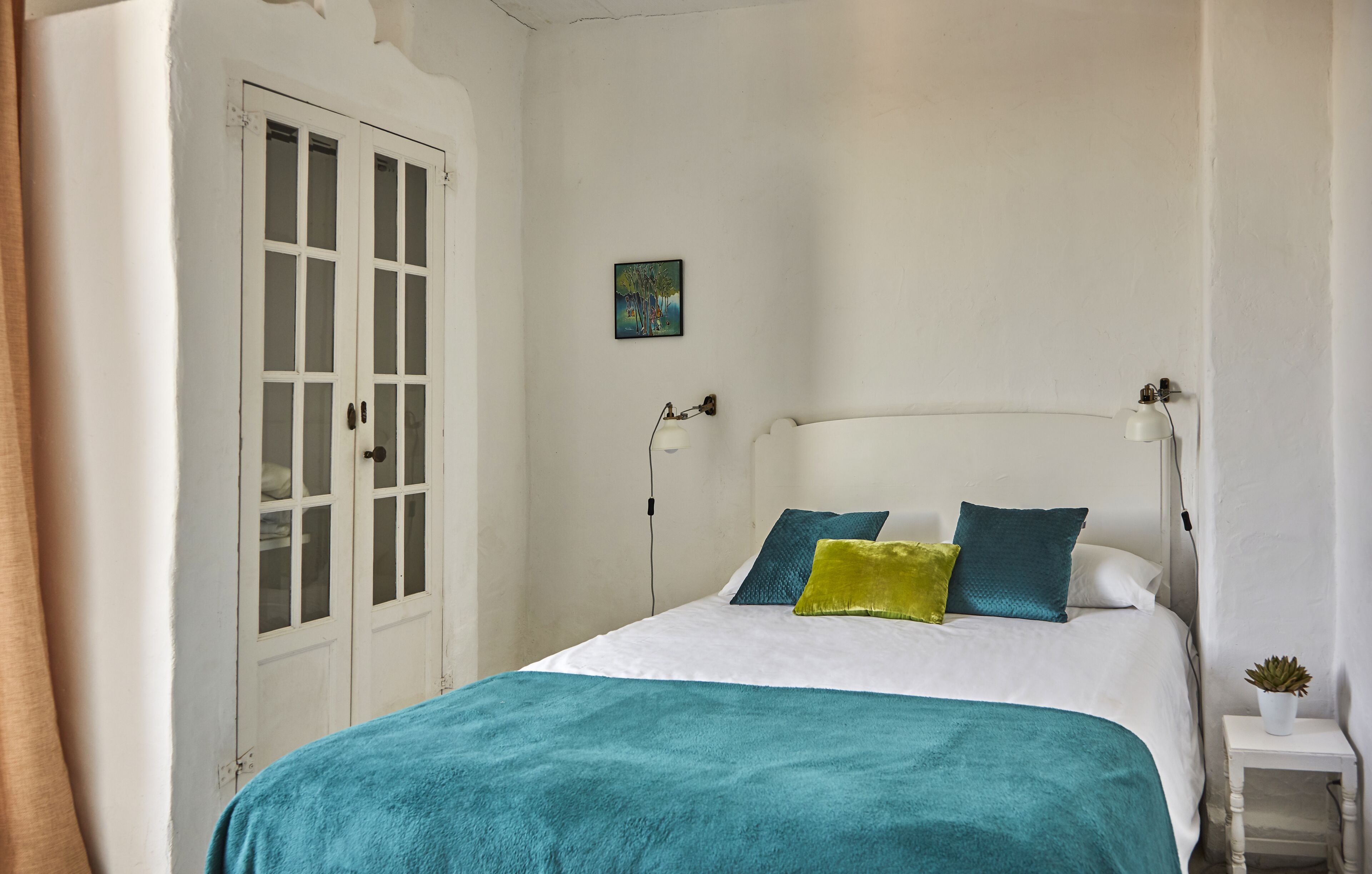 A bedroom with a bed, white walls, a door, a small table with a plant, and a painting on the wall.