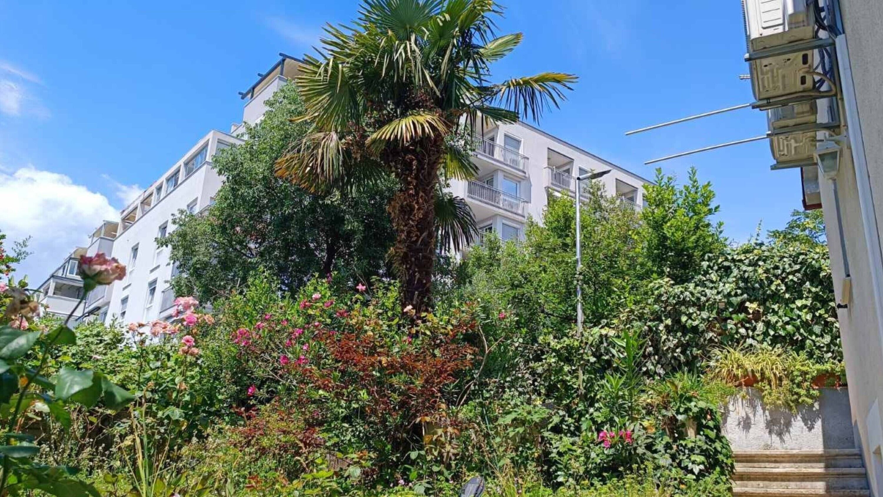 Ein mehrstöckiges Gebäude mit Balkonen, umgeben von üppiger Vegetation und blühenden Pflanzen.
