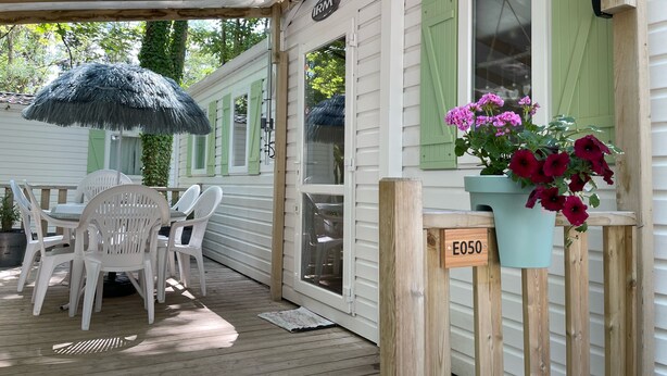 Ein überdachter Außenbereich mit Holzterrasse, weißen Plastikstühlen, einem Tisch und einem Blumentopf mit rosa Blüten.