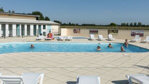Ein Schwimmbecken mit darin badenden Personen, umgeben von Liegestühlen und einem Gebäude mit einem Ziegeldach.