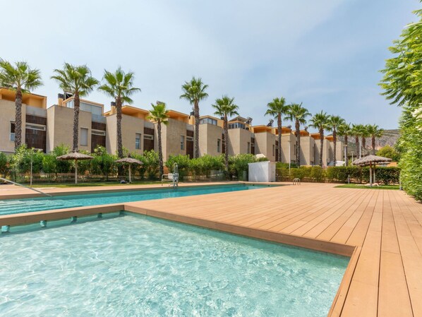 Zona de piscina con terraza de madera, palmeras y un edificio con balcones.