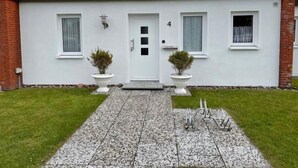 Een modern huis met twee verdiepingen, een witte gevel, een bakstenen schoorsteen en het getal '4' op de voordeur. Aan weerszijden van de ingang staan twee potplanten.