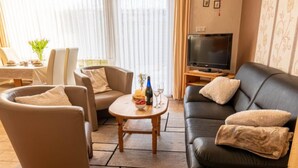 Een woonkamer met een bank, twee fauteuils, een salontafel, een televisie en een eethoek met een tafel en stoelen.