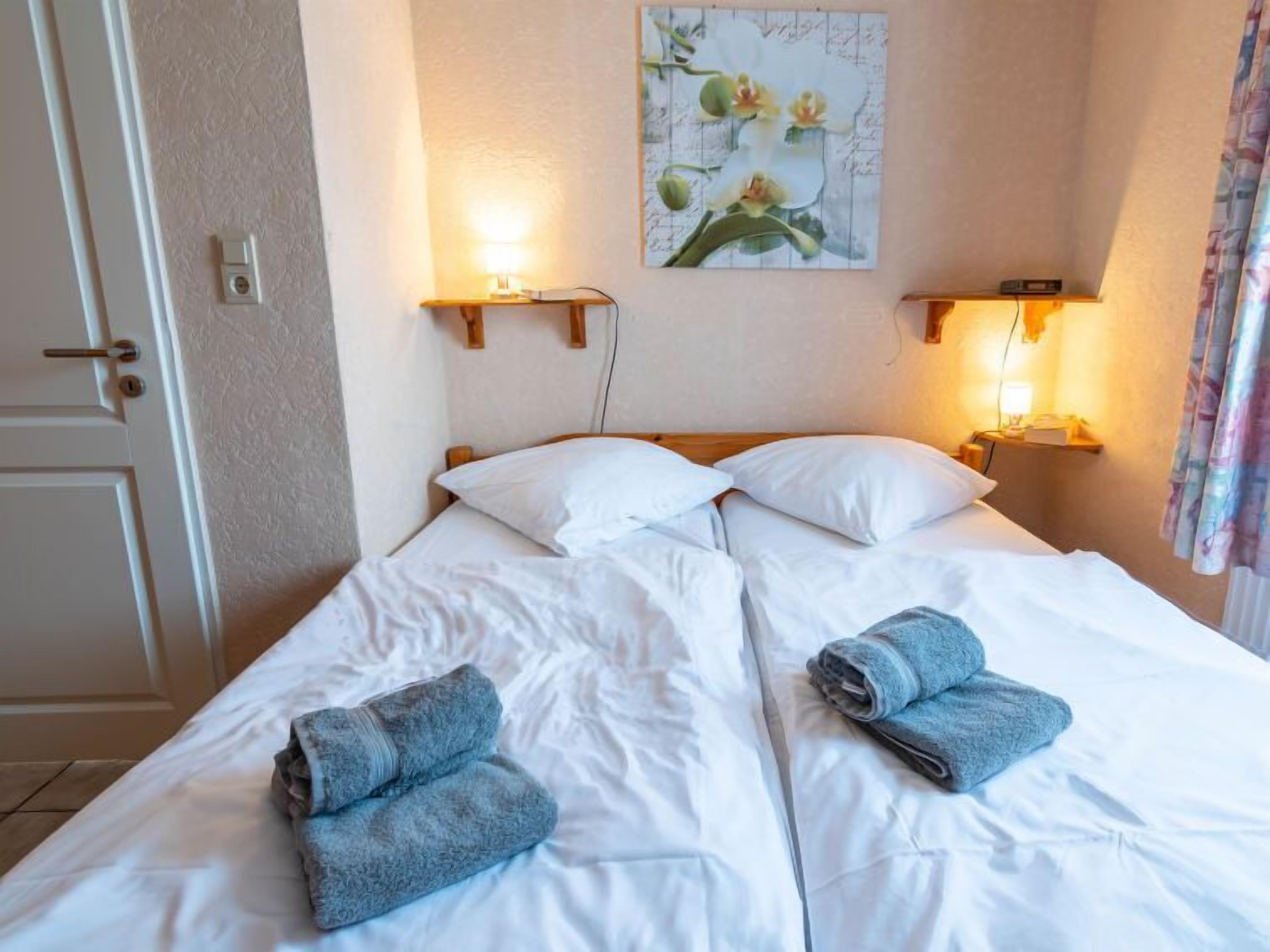 Ein Doppelbett mit weißen Bettwäsche und zwei gefalteten Handtüchern, Nachttischlampen, ein an der Wand hängendes Blumenbild und eine Tür mit Griff.
