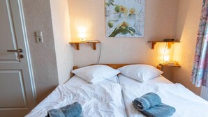 Een tweepersoonsbed met witte lakens en twee opgevouwen handdoeken, bedlampen, een aan de muur hangend schilderij met bloemen, en een deur met een hendel.