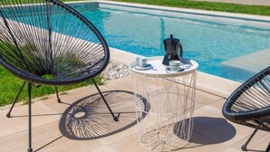 Un espace aménagé au bord de la piscine, avec deux chaises au design épuré en fil de fer noir, une petite table ronde sur laquelle se trouvent une cafetière et des tasses, et une piscine à l’eau d’un bleu limpide.