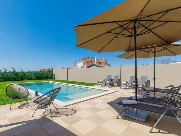Une piscine entourée d’une clôture et de verdure, avec des chaises longues et des parasols.