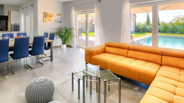 Un salon moderne avec un grand canapé orange, un coin repas avec des chaises bleues et une vue sur une piscine, visible à travers de grandes fenêtres.