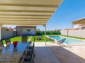 Une terrasse couverte avec une piscine, un coin repas avec des chaises et un petit jardin.