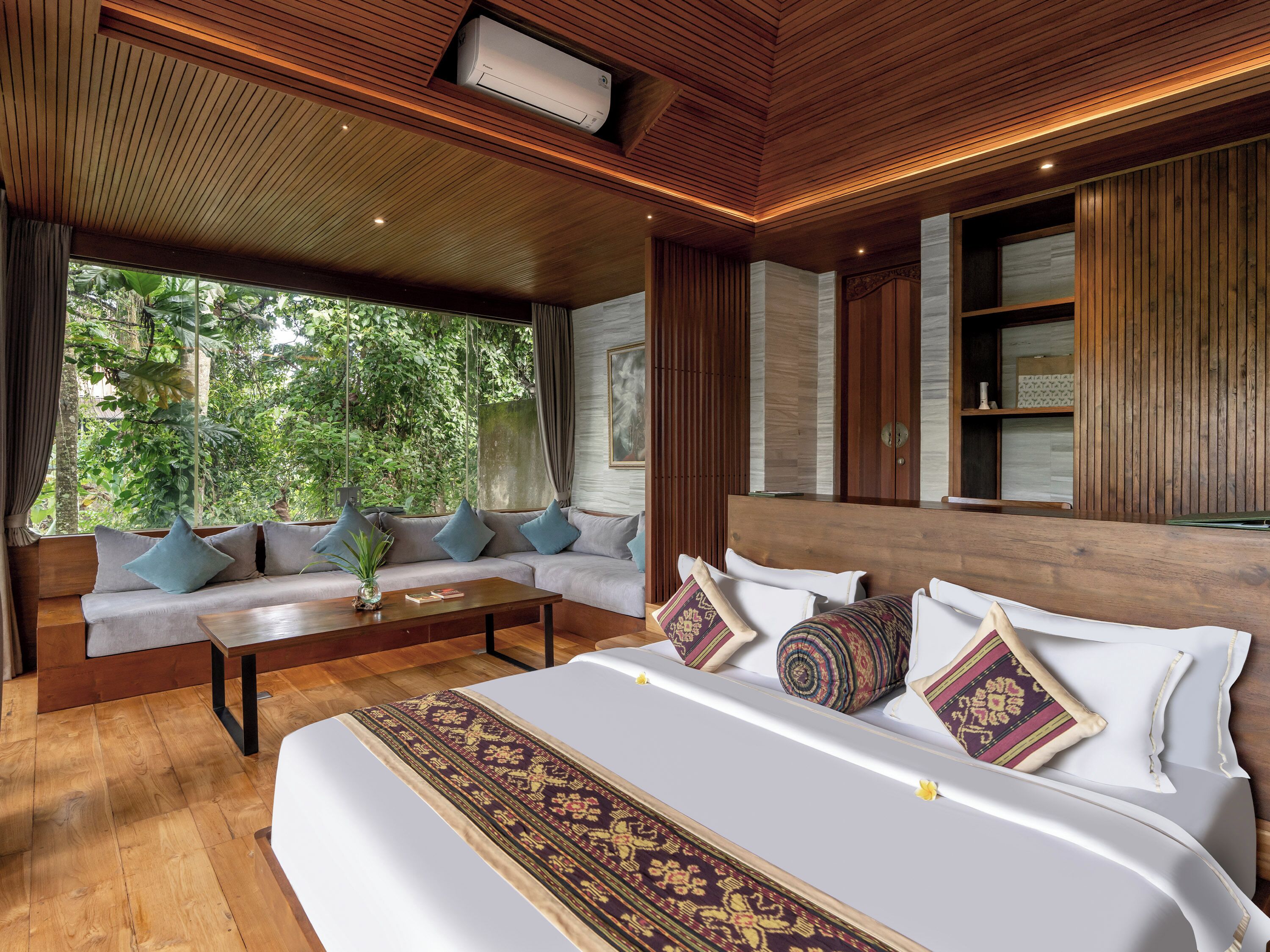 A modern bedroom with a large bed, a sofa, a wooden ceiling, and a view of greenery outside.
