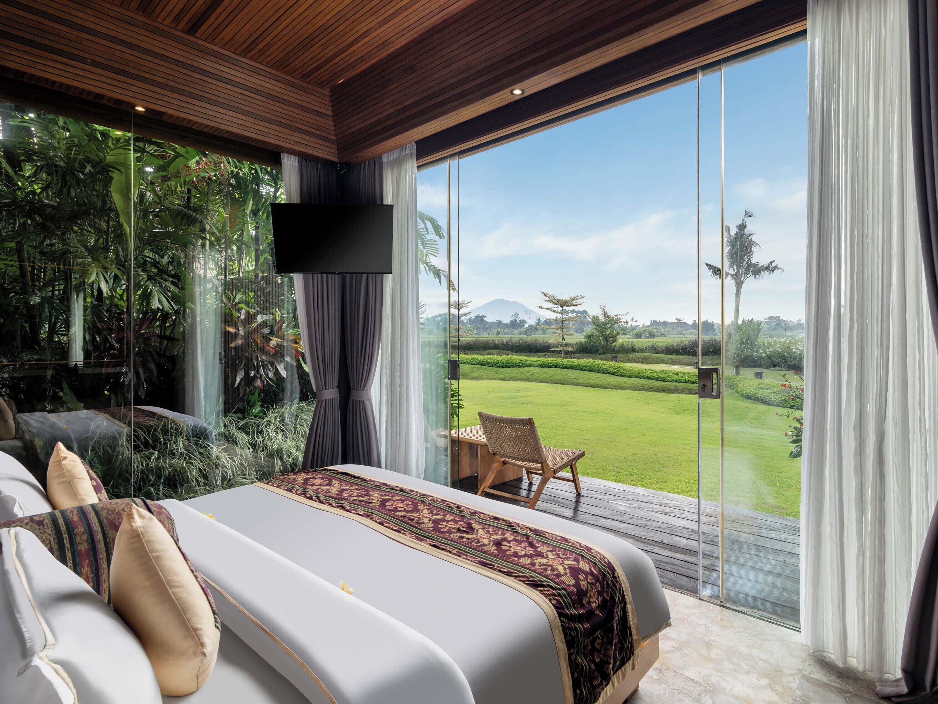 A bedroom with a large bed, a TV, and a view of a green landscape through a glass door.