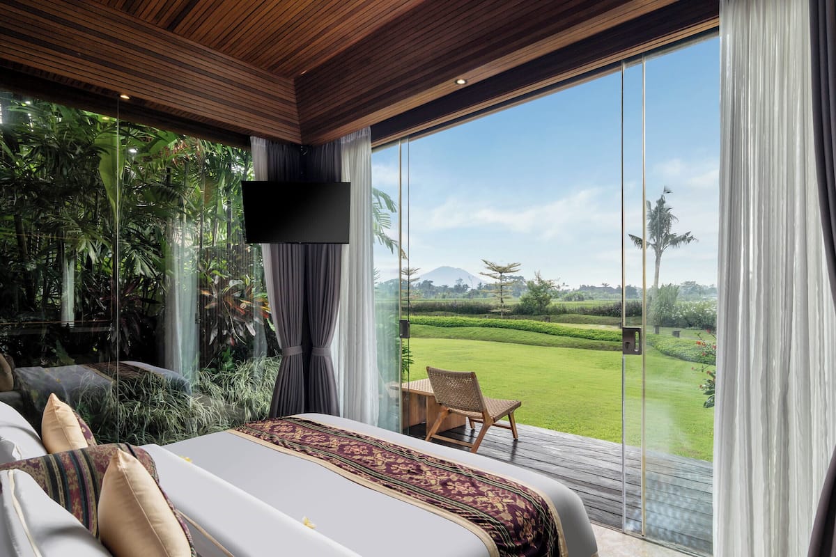 A bedroom with a large bed, a TV, and a view of a green landscape through a glass door.