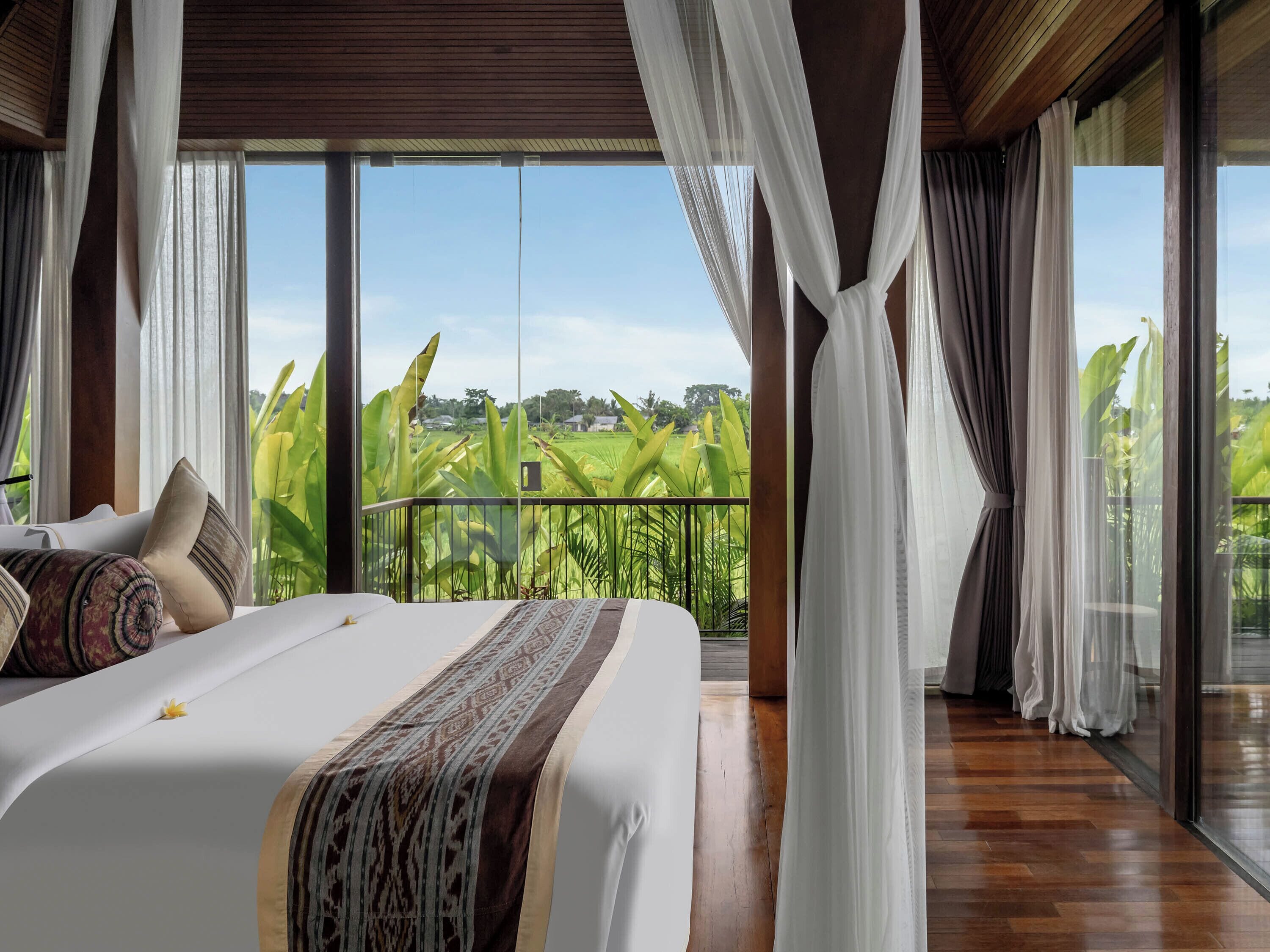 A bedroom with a large bed, sheer curtains, and a view of a green landscape.