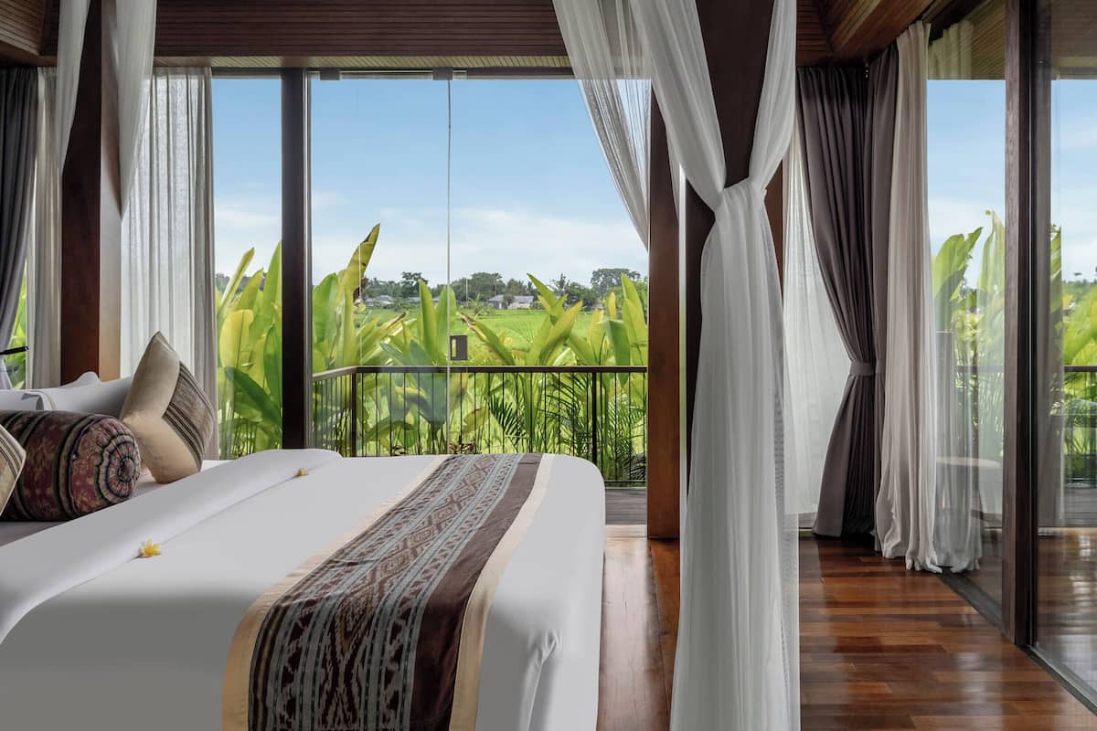A bedroom with a large bed, sheer curtains, and a view of a green landscape.