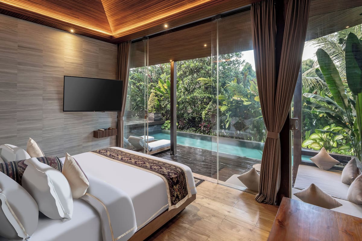 A modern bedroom with a large bed, a TV, and a view of a lush garden and pool.