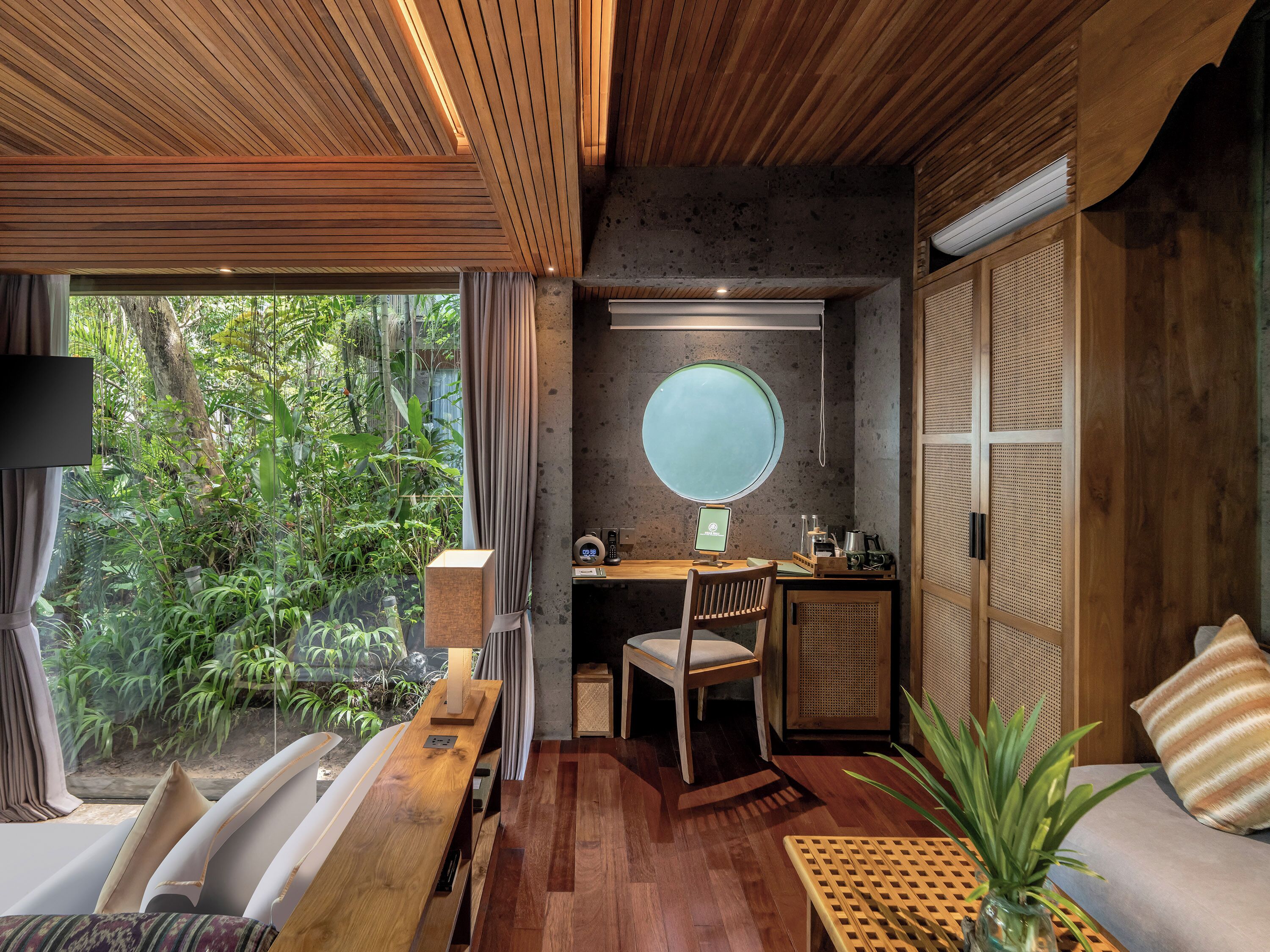 A modern room with a wooden ceiling, a round window, a desk with a computer, a chair, a sofa, and a view of greenery outside.