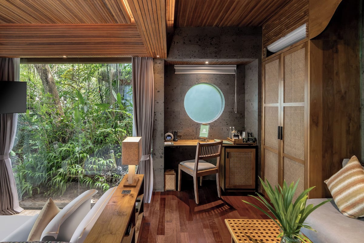 A modern room with a wooden ceiling, a round window, a desk with a computer, a chair, a sofa, and a view of greenery outside.