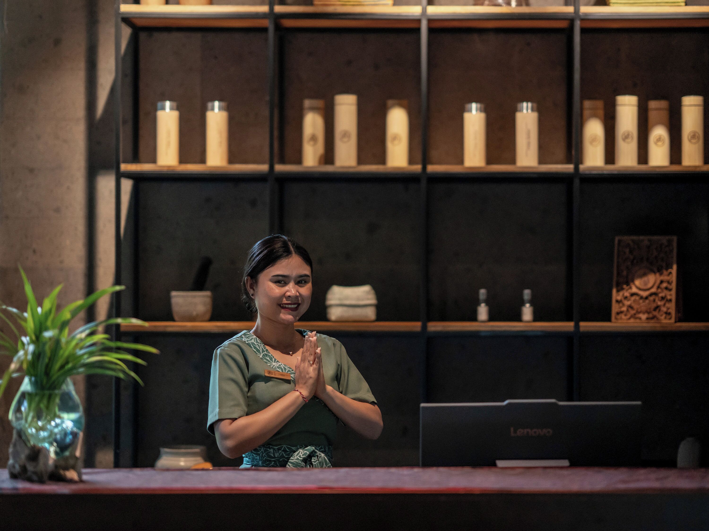 A reception desk with a laptop, a potted plant, and a shelf with bottles and a framed picture.