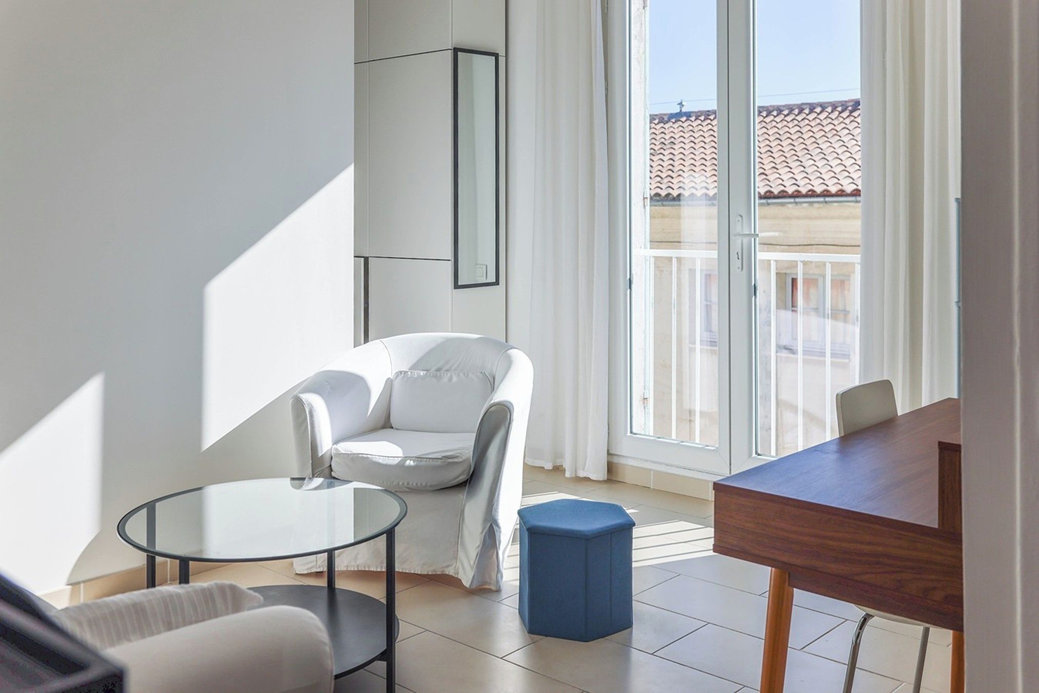 A modern living room with a white armchair, a glass coffee table, a blue ottoman, and a wooden dining table.