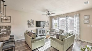 A spacious living room with a high ceiling, a ceiling fan, and a large window with patterned curtains. There is a flat-screen TV mounted on the wall, a wooden cabinet, and a dining area with a bar stool and a glass-top table. - Brand-New Townhome Near UNT/TWU & The Square! (Denton)