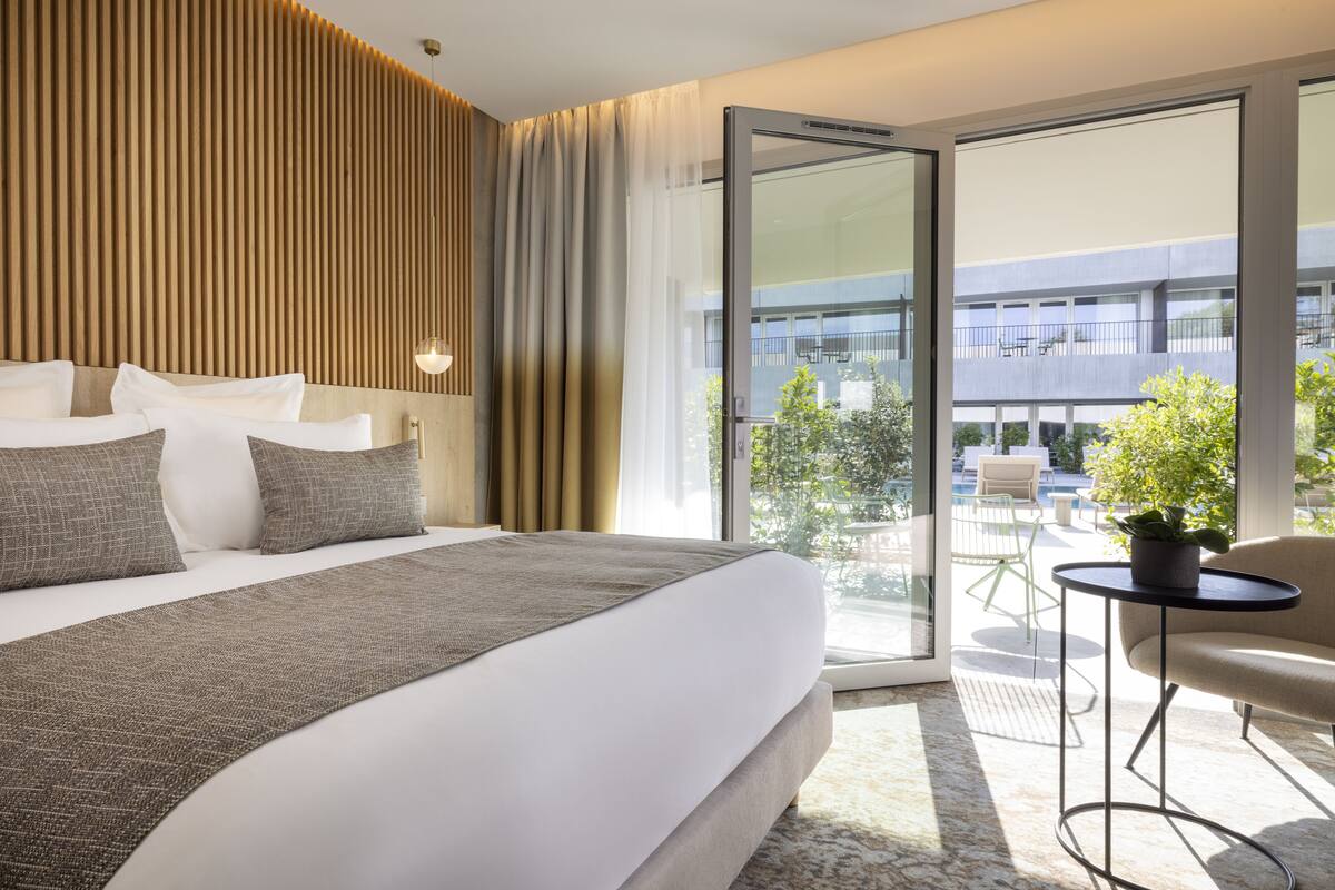 A modern hotel room with a large bed, a small table with a plant, and a balcony with outdoor seating.