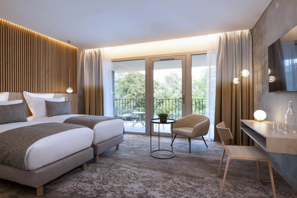 A modern hotel room with a large bed, two bedside tables, a seating area with chairs, a small table with a plant, and a TV mounted on the wall.