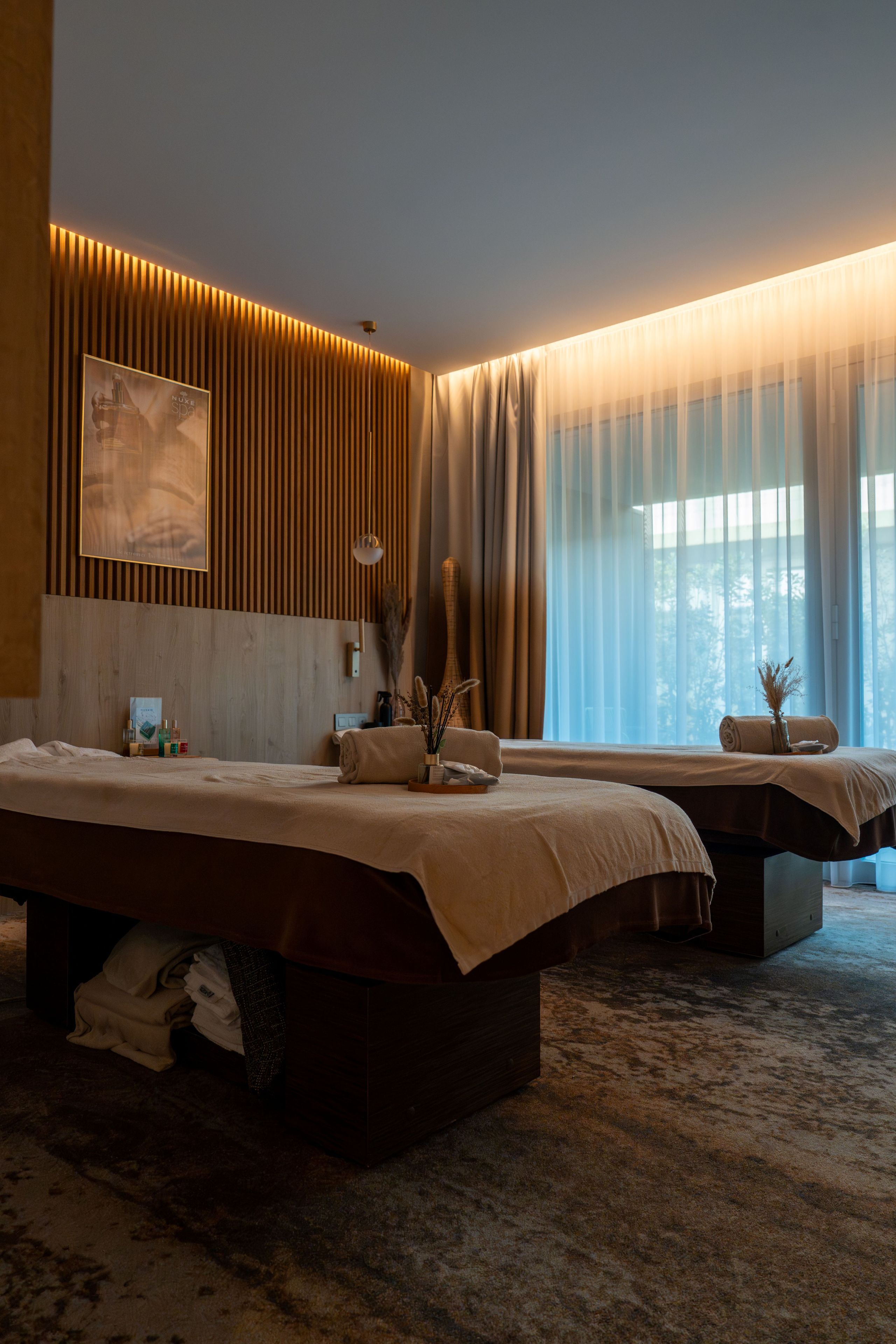 A hotel room with two beds, a large window with sheer curtains, a wall-mounted light, and a framed picture.