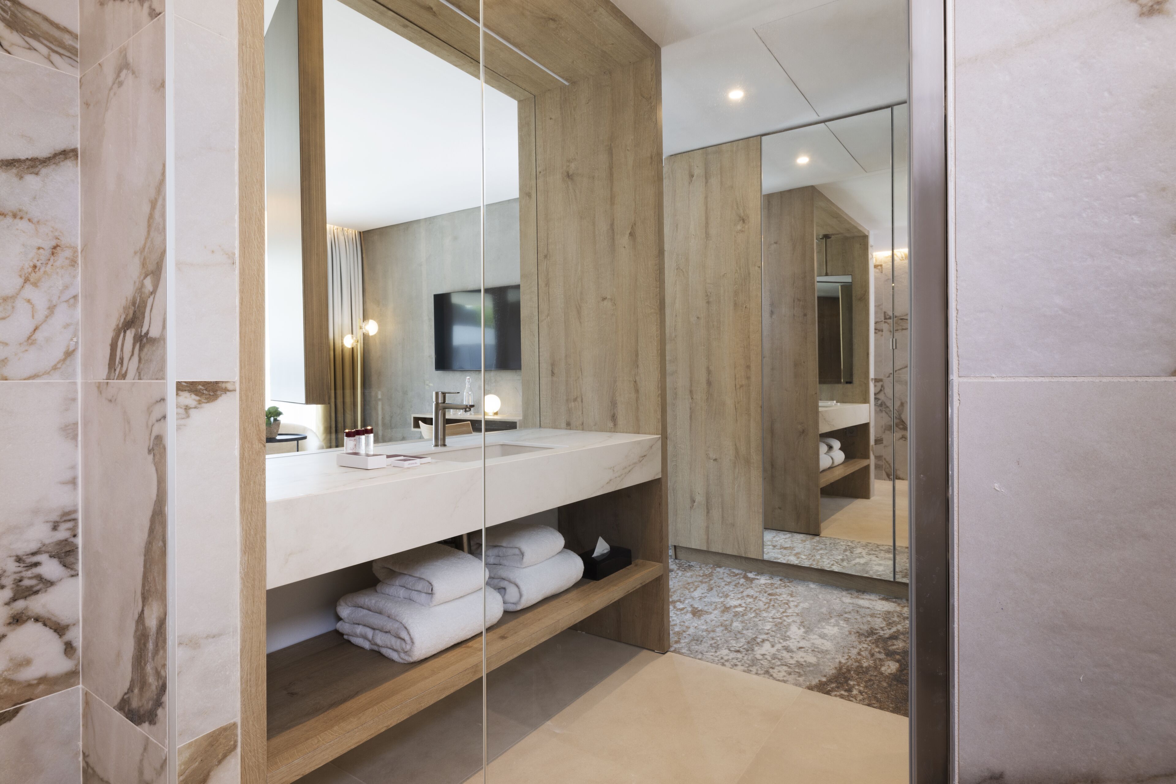 A modern bathroom with a large mirror, marble tiles, wooden shelves, and a television.