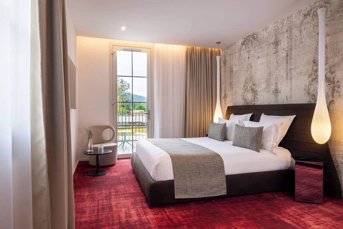 A hotel room with a large bed, a small round table, a chair, and a view of the outdoors through a window.