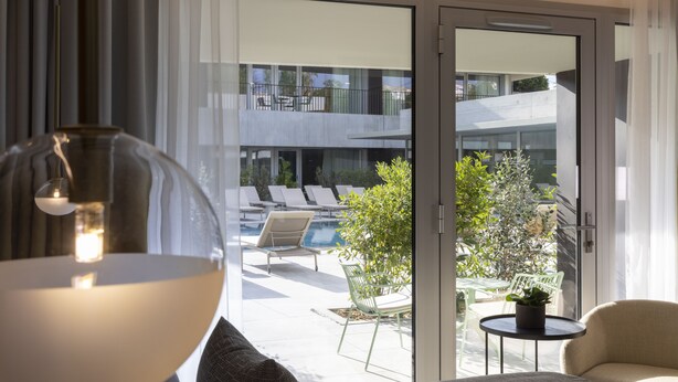 Una habitación de hotel moderna con un ventanal grande, una cama, una mesa redonda con una planta y vistas a la piscina exterior y al balcón.