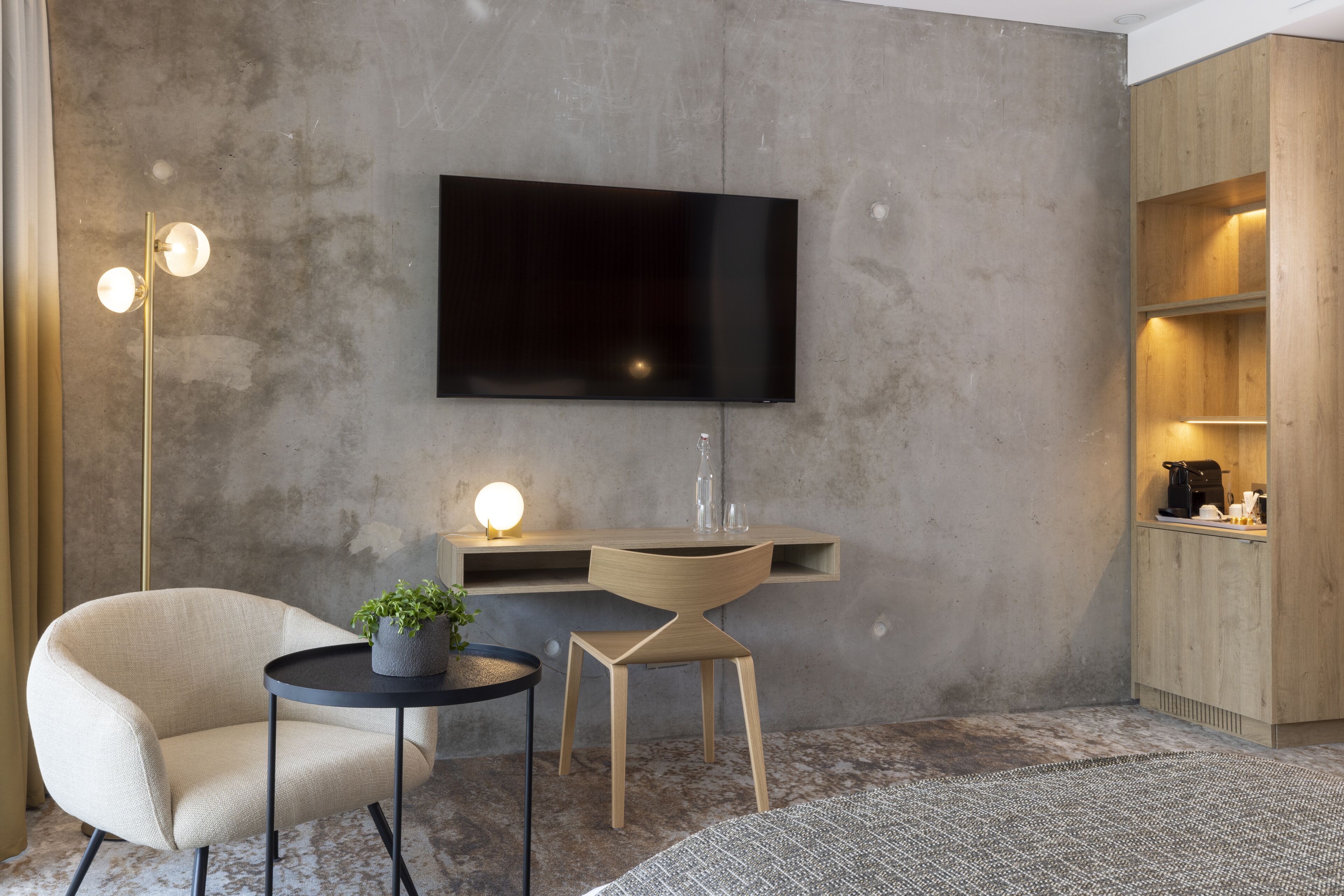 Una sala de estar moderna con un televisor de pantalla plana montado en una pared de concreto, una mesa redonda de centro con una planta en maceta, un sillón color beige y una pequeña mesita de noche con una lámpara.