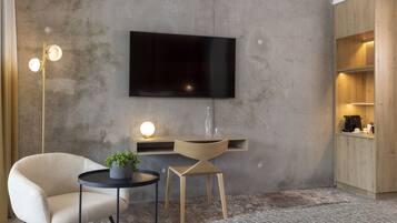 Una sala de estar moderna con un televisor de pantalla plana montado en una pared de concreto, una mesa redonda de centro con una planta en maceta, un sillón color beige y una pequeña mesita de noche con una lámpara.