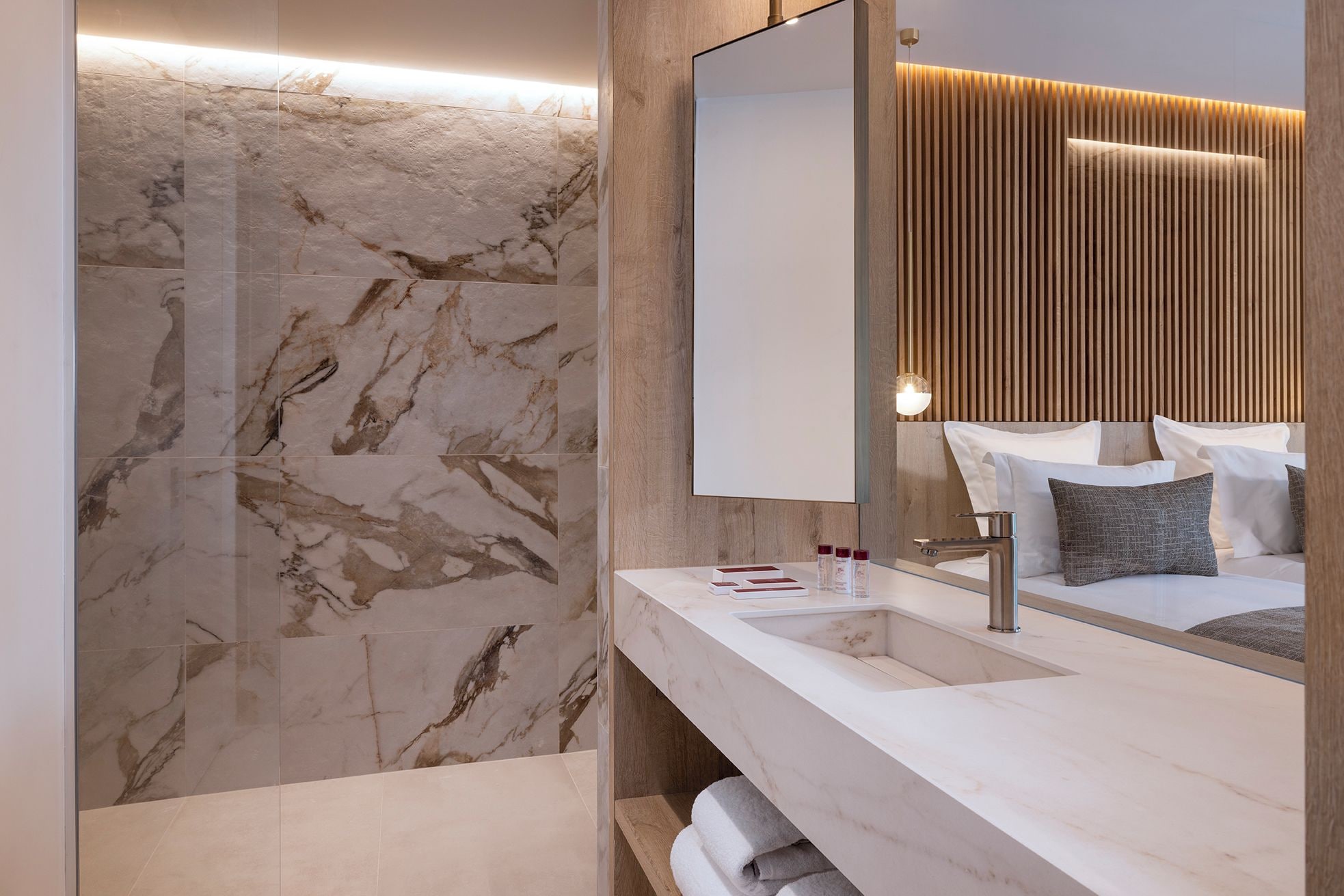 Une salle de bain moderne avec un mur en marbre, un grand miroir, un mur recouvert de panneaux en bois et un lavabo blanc agrémenté d’accessoires de toilette.