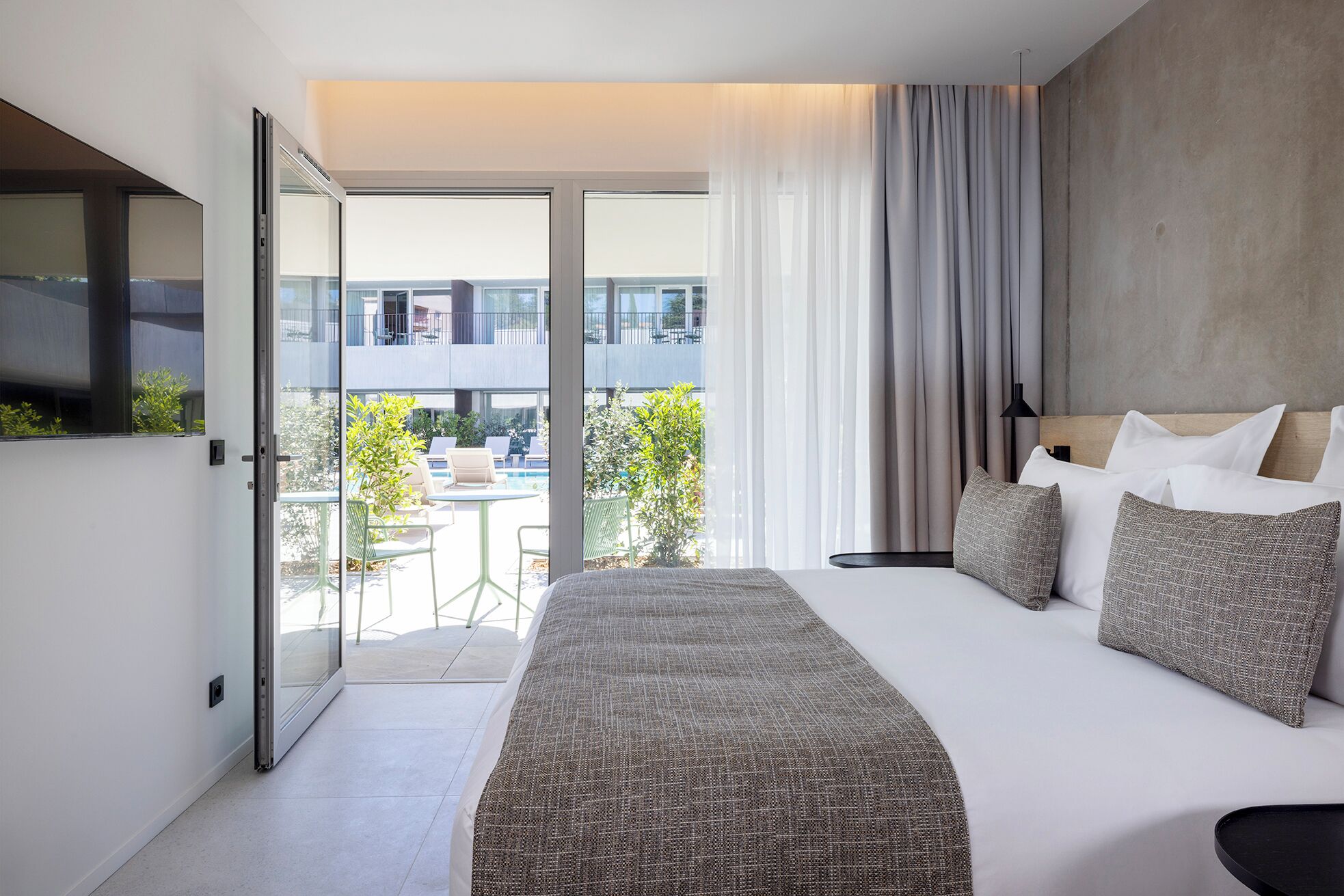 A modern hotel room with a bed, a TV, a balcony with a pool, and a view of the city.