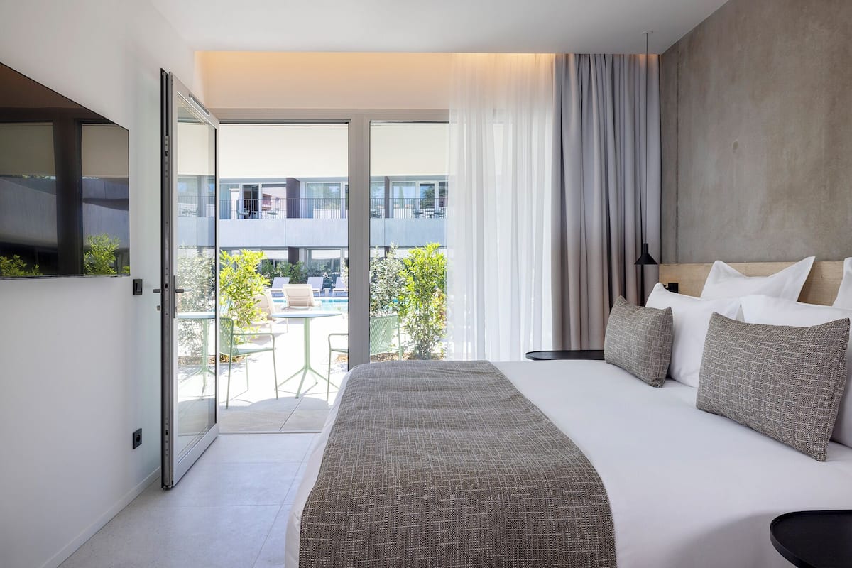 A modern hotel room with a bed, a TV, a balcony with a pool, and a view of the city.