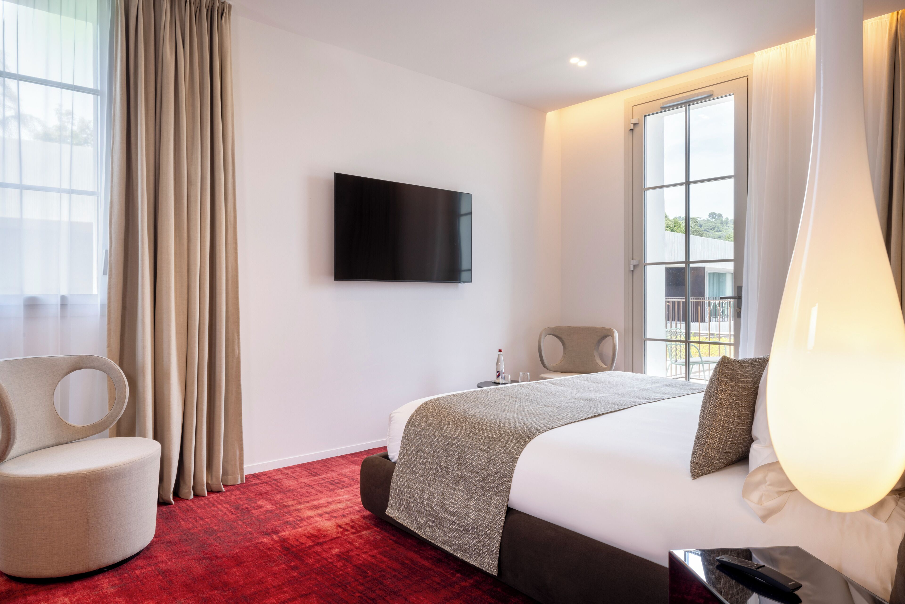 Una habitación de hotel moderna con una cama grande, un televisor de pantalla plana, una alfombra roja y vistas al exterior.