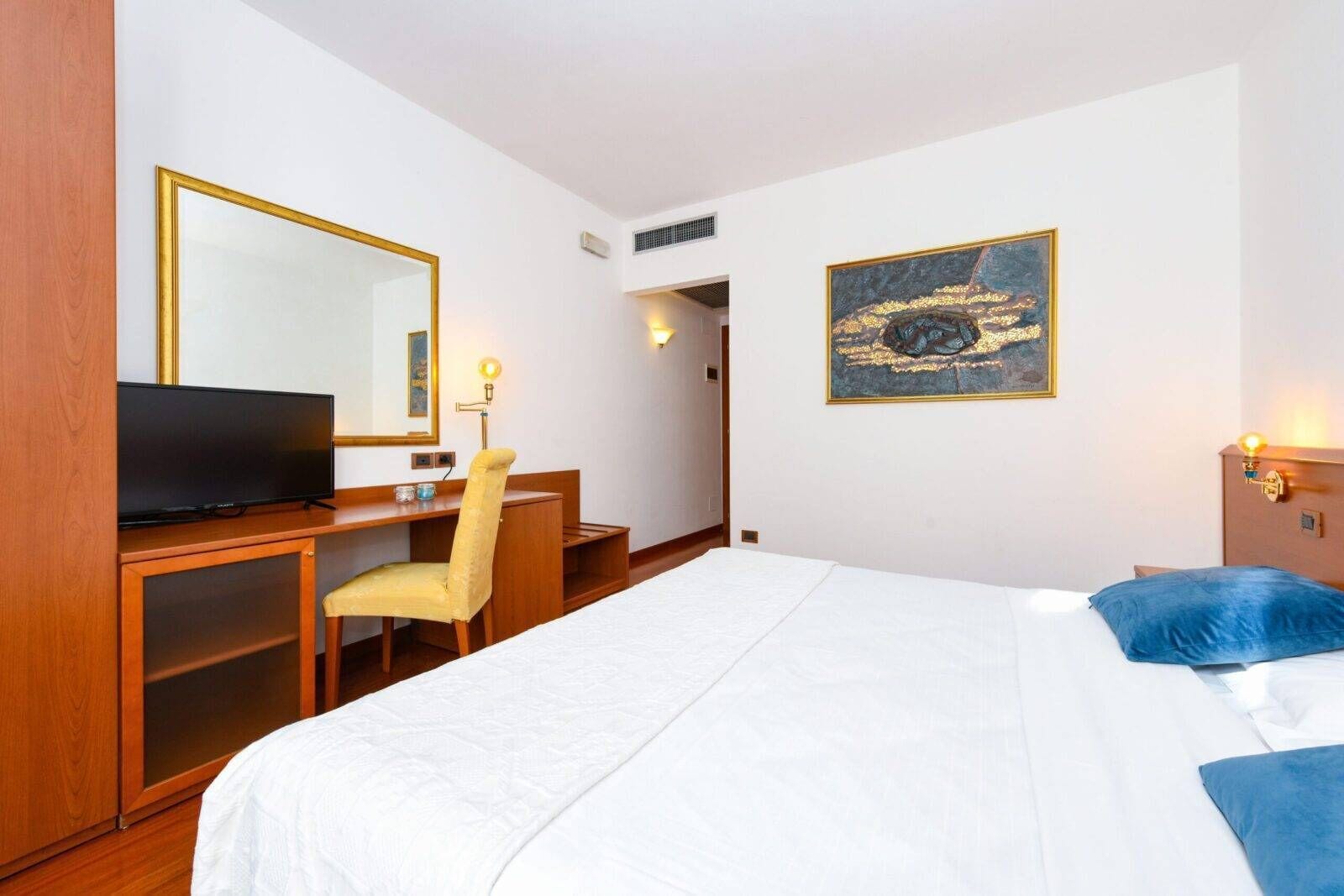 Habitación de hotel con cama, un escritorio con silla, televisor, un cuadro en la pared y una puerta que da a otra habitación.
